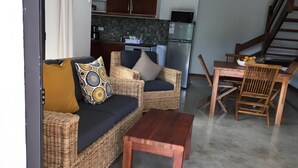Ein Wohnzimmer mit einer Couch, zwei Sesseln, einem Holztisch und einem Esstisch mit Stühlen. Im Hintergrund ist eine Küche mit Schränken und Geräten zu sehen.