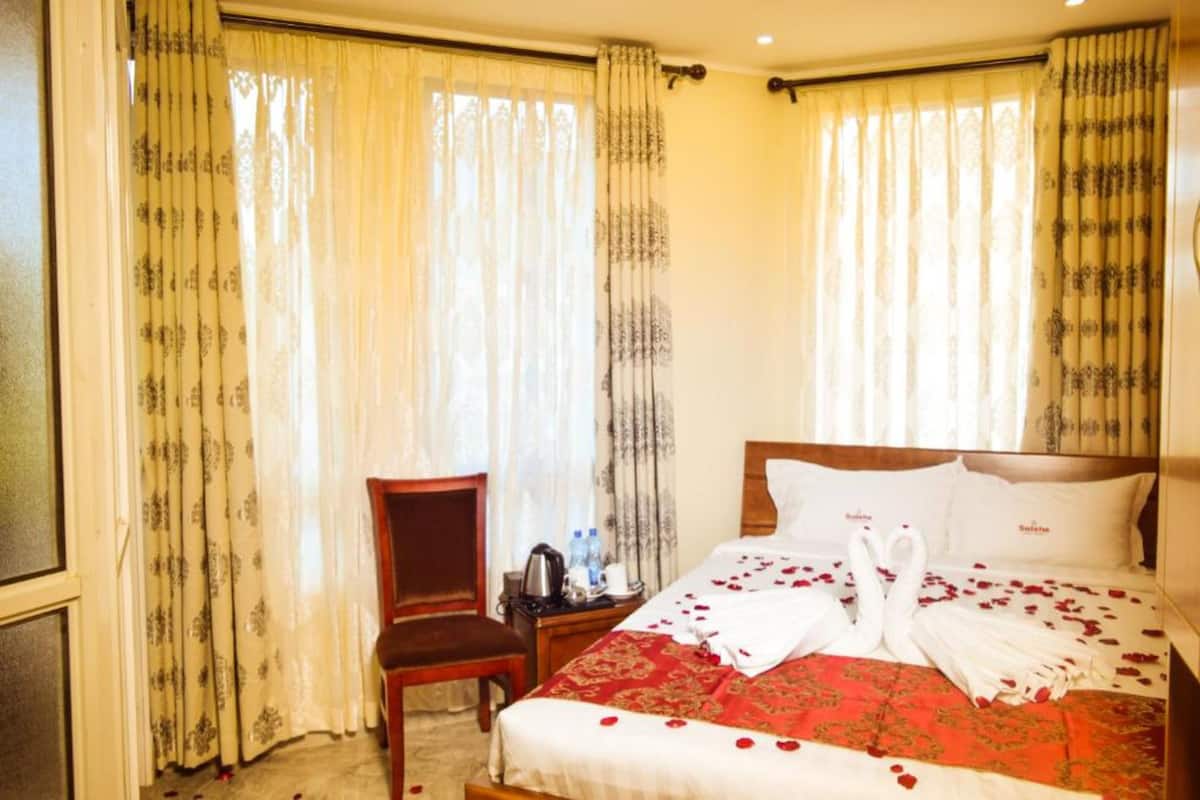 A hotel room with a bed, curtains, a chair, and a small table with a kettle and water bottles.