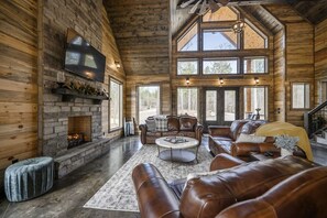 Interior espacioso de una cabaña de troncos con chimenea, mobiliario de cuero y amplias ventanas que ofrecen vistas al exterior.