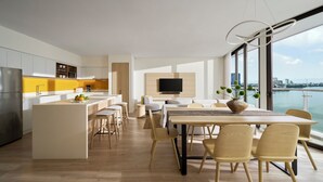 Una cocina moderna con una isla central, un área de comedor con mesa y sillas, y un amplio ventanal con vistas a la ciudad y al agua.