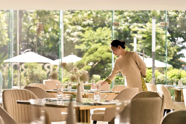 A restaurant with tables set for dining, a waiter attending to guests, and a view of greenery through large windows. - PARKROYAL Serviced Suites Hanoi (Hanoi)