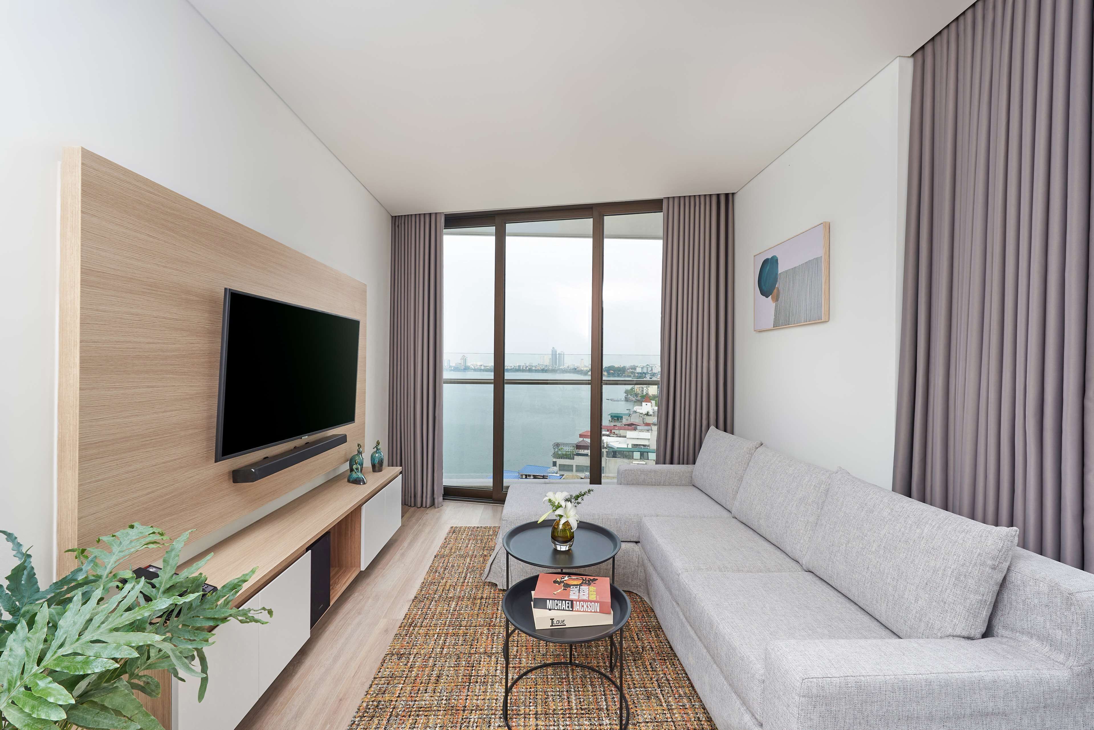 A modern living room with a flat-screen TV, a grey sofa, a small table with a book, and a view of the city through large windows.