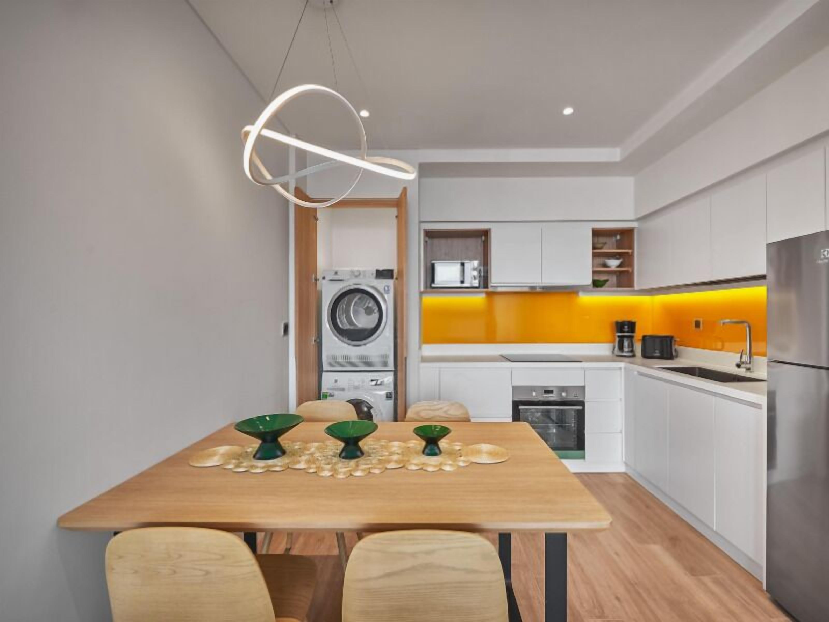 A modern kitchen with a built-in washer and dryer, a dining area with a wooden table and chairs, and a bright yellow backsplash.