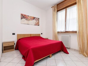 Un dormitorio con cama, mesita de noche, radiador y ventana con cortinas.