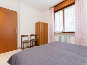 Une chambre à coucher comprenant un lit, deux chaises, une armoire, une fenêtre avec des rideaux et un radiateur.