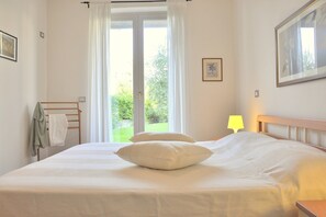 Una camera da letto con un letto, un comodino, una lampada, una finestra con tende e vista su del verde all'esterno.