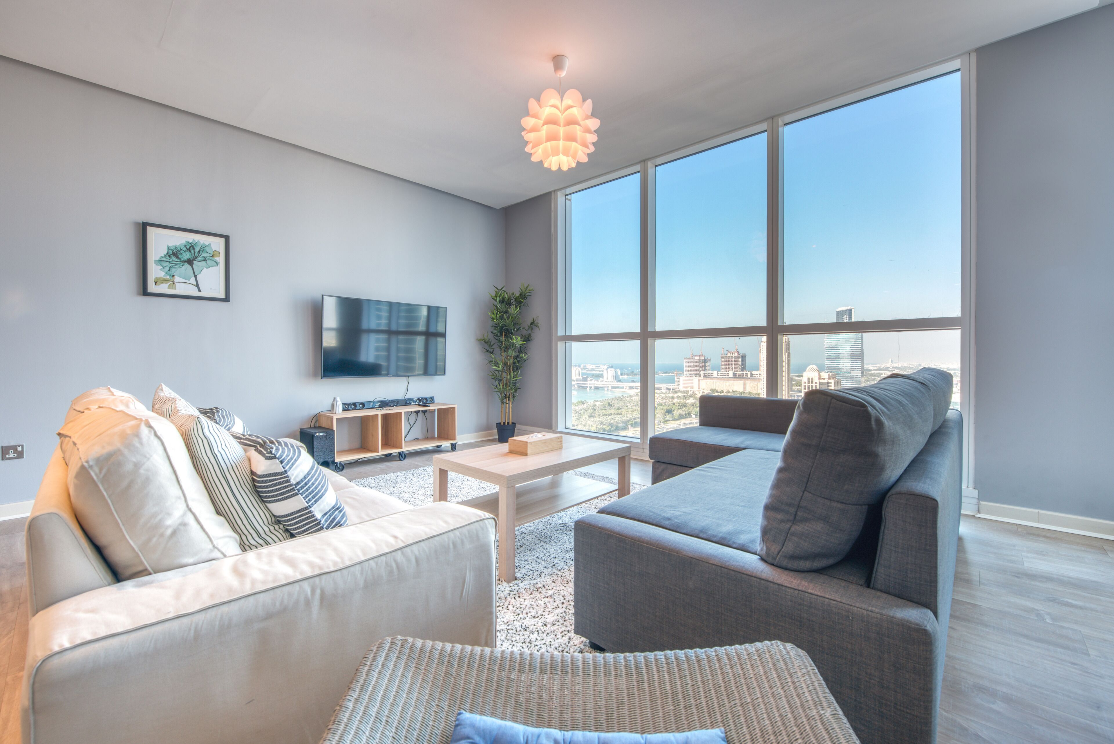 Ein modernes Wohnzimmer mit grauem Sofa, grauem Sessel, einem kleinen Couchtisch und einem Fernseher an der Wand. Ein großes Fenster bietet Blick auf die Stadt.