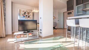 Una sala moderna con un televisor de pantalla plana, una mesa de centro de vidrio y un comedor con sillas blancas, con una cocina al fondo.