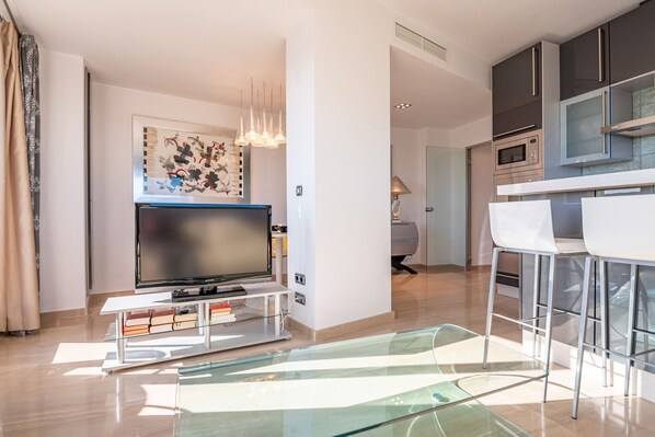 A modern living room with a flat-screen TV, a glass coffee table, and a dining area with white chairs and a kitchen in the background. - Apartamentos Cardenio Penthouse PLUS - Ayamonte Centre AEHCA3P01 (Ayamonte)