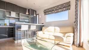 Una sala de estar moderna con una mesa de centro de vidrio, un sofá color beige y una zona de cocina con armarios oscuros y una ventana con cortinas estampadas.