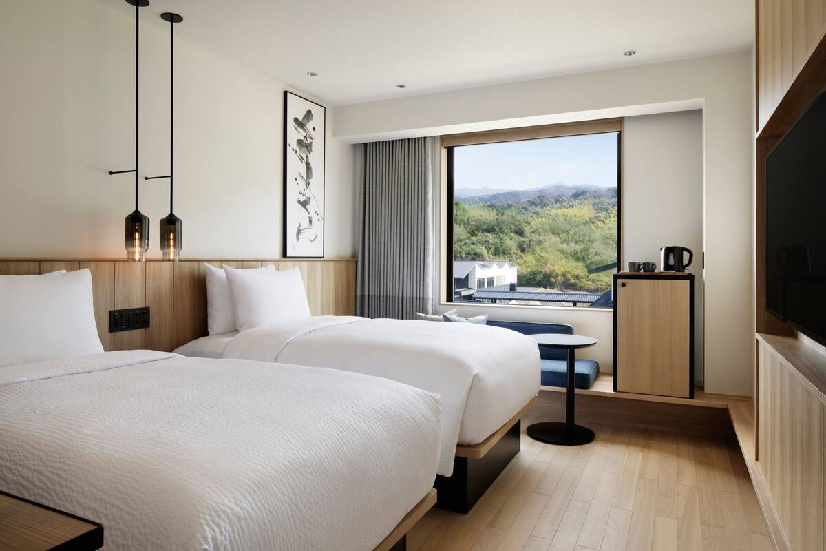 Habitación de hotel con dos camas, un ventanal con vistas a un bosque, una mesita con una silla y un televisor en la pared.