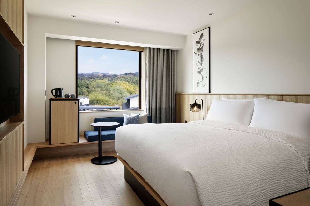 Una habitación de hotel moderna con una cama grande, un pequeño espacio de descanso, un ventanal con vistas a zonas verdes y un televisor de pantalla plana.