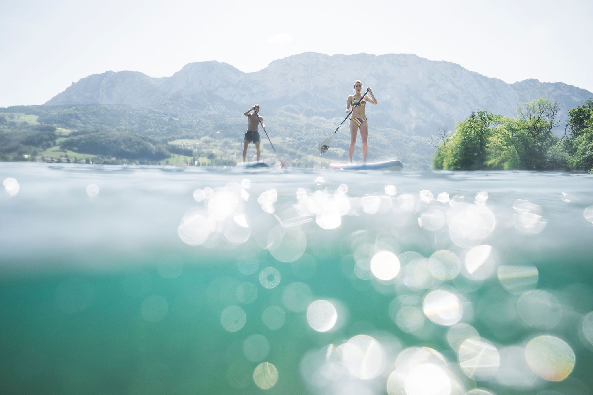 Two people paddleboarding on a calm lake with mountains in the background.