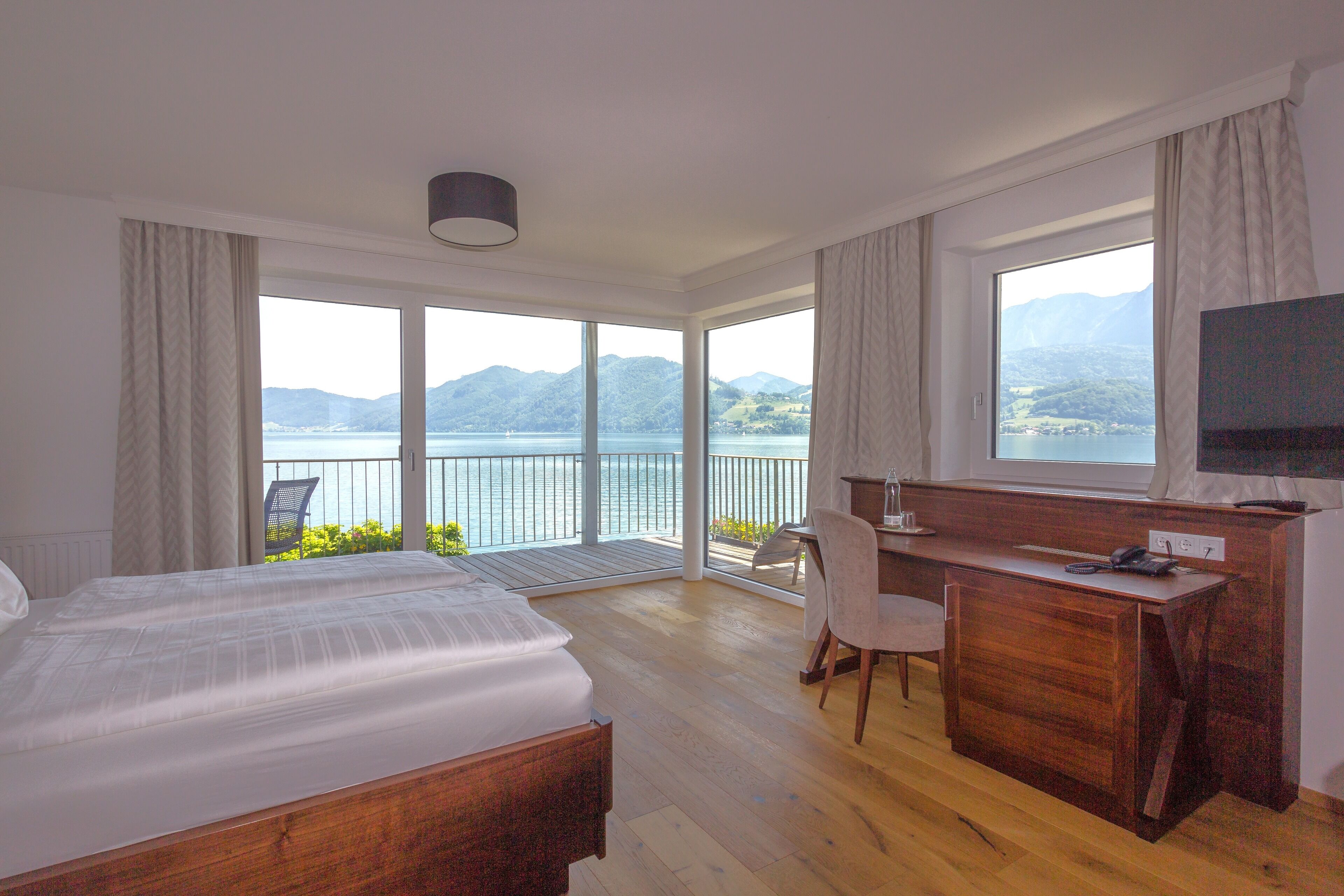 A hotel room with a large bed, a desk, and a TV. It has a balcony with a view of the mountains and lake.