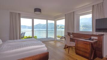 A hotel room with a large bed, a desk, and a TV. It has a balcony with a view of the mountains and lake.
