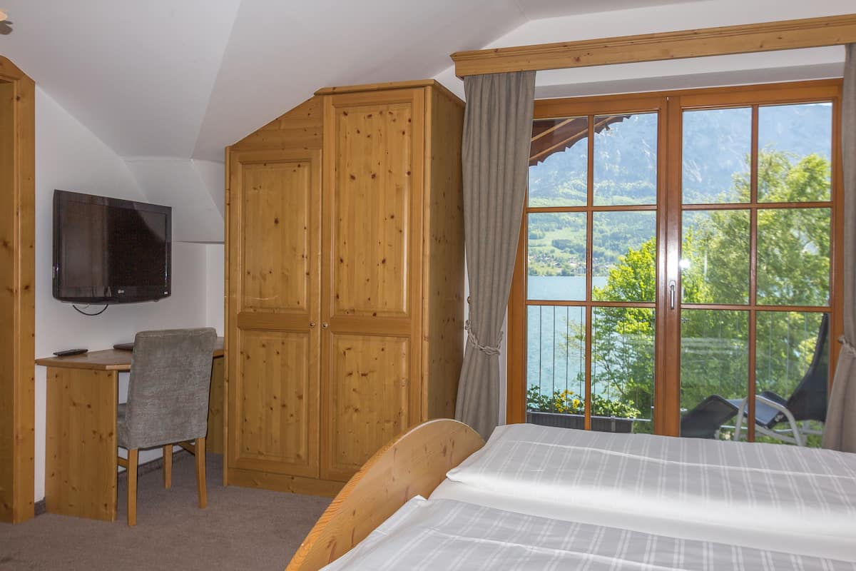 Une chambre à coucher comprenant un lit, un bureau, une chaise, une télévision et une grande fenêtre donnant sur un lac et des montagnes.