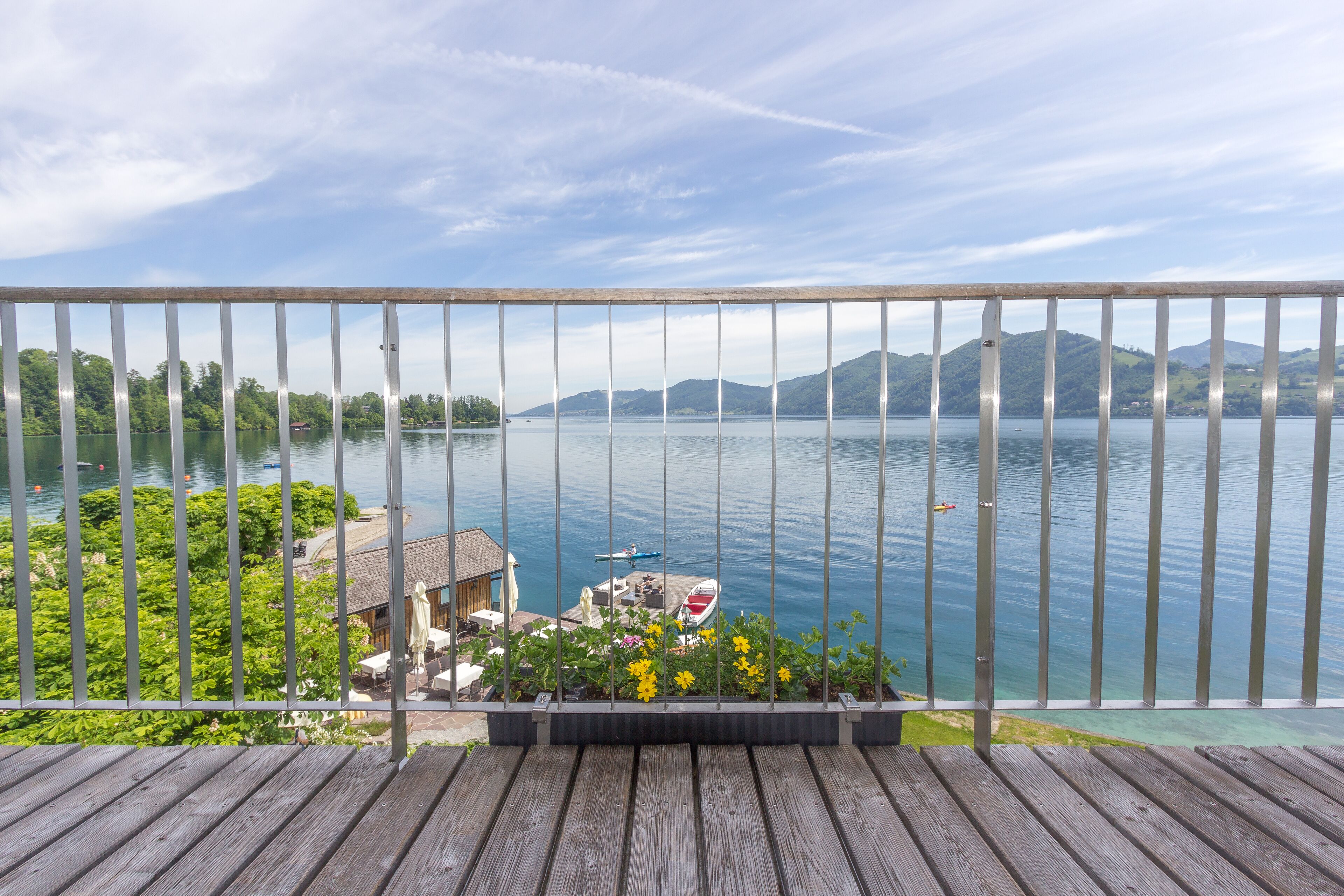 A wooden deck with a metal railing overlooking a lake, a boat docked near the shore, and a building with outdoor seating.