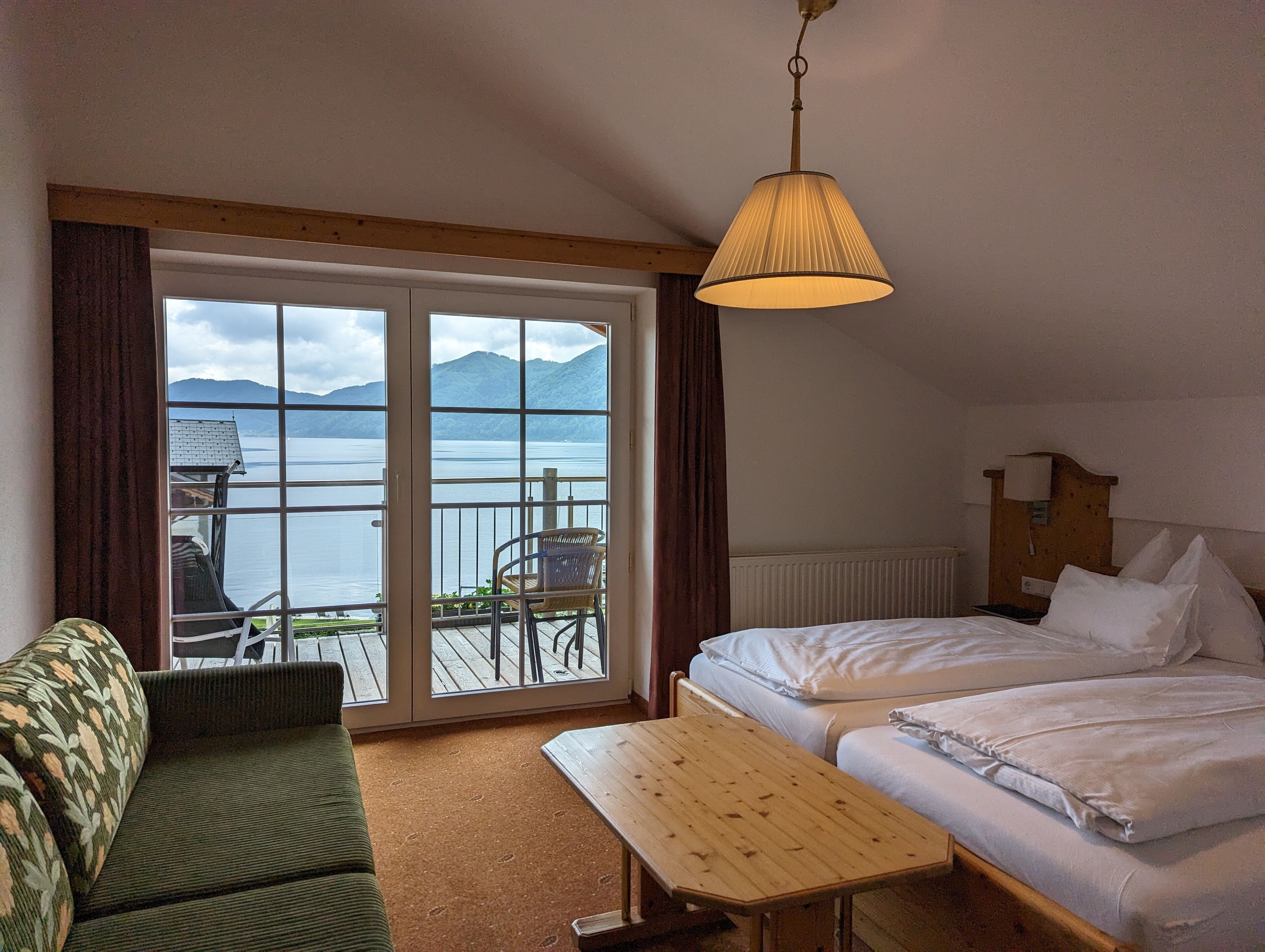 Habitación de hotel con cama, una mesita, un sofá y vistas a un lago y montañas a través de amplias ventanas.