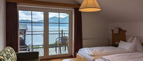 Habitación de hotel con cama, una mesita, un sofá y vistas a un lago y montañas a través de amplias ventanas.