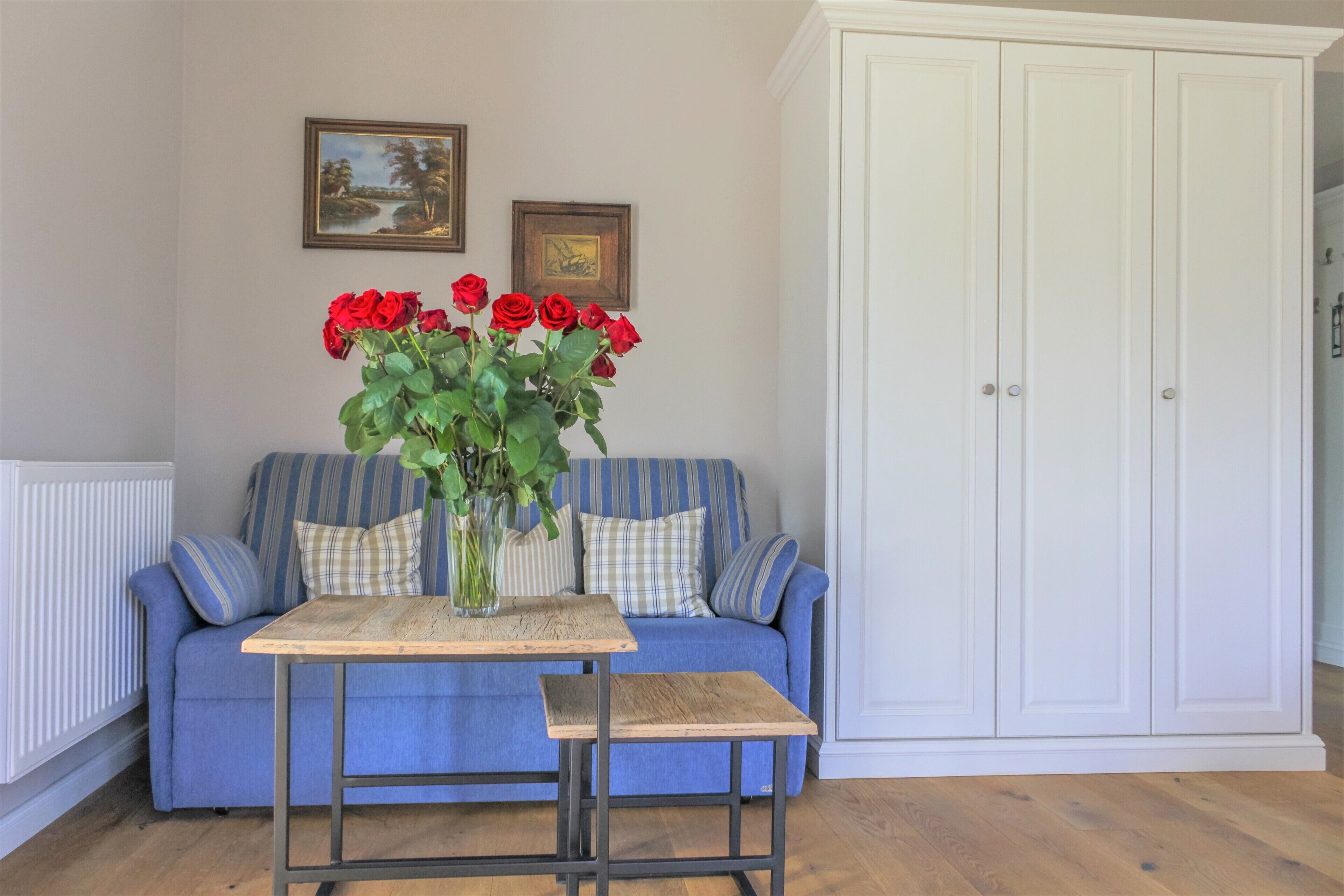 Une pièce avec un canapé bleu, une petite table en bois sur laquelle est posé un vase de roses rouges, et une armoire blanche.