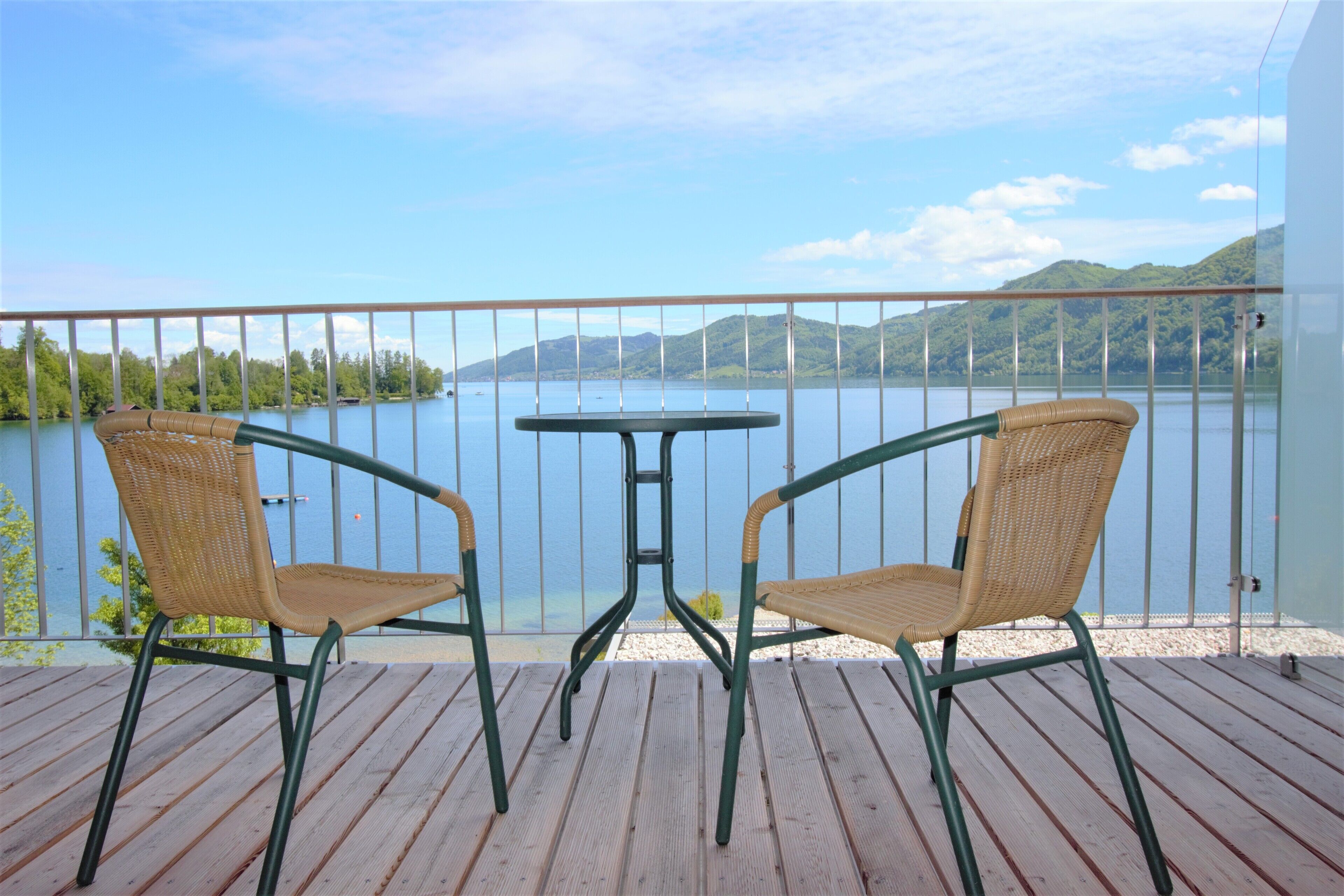 Una terraza de madera con dos sillas de mimbre y una mesita, con vistas a un lago y montañas al fondo.