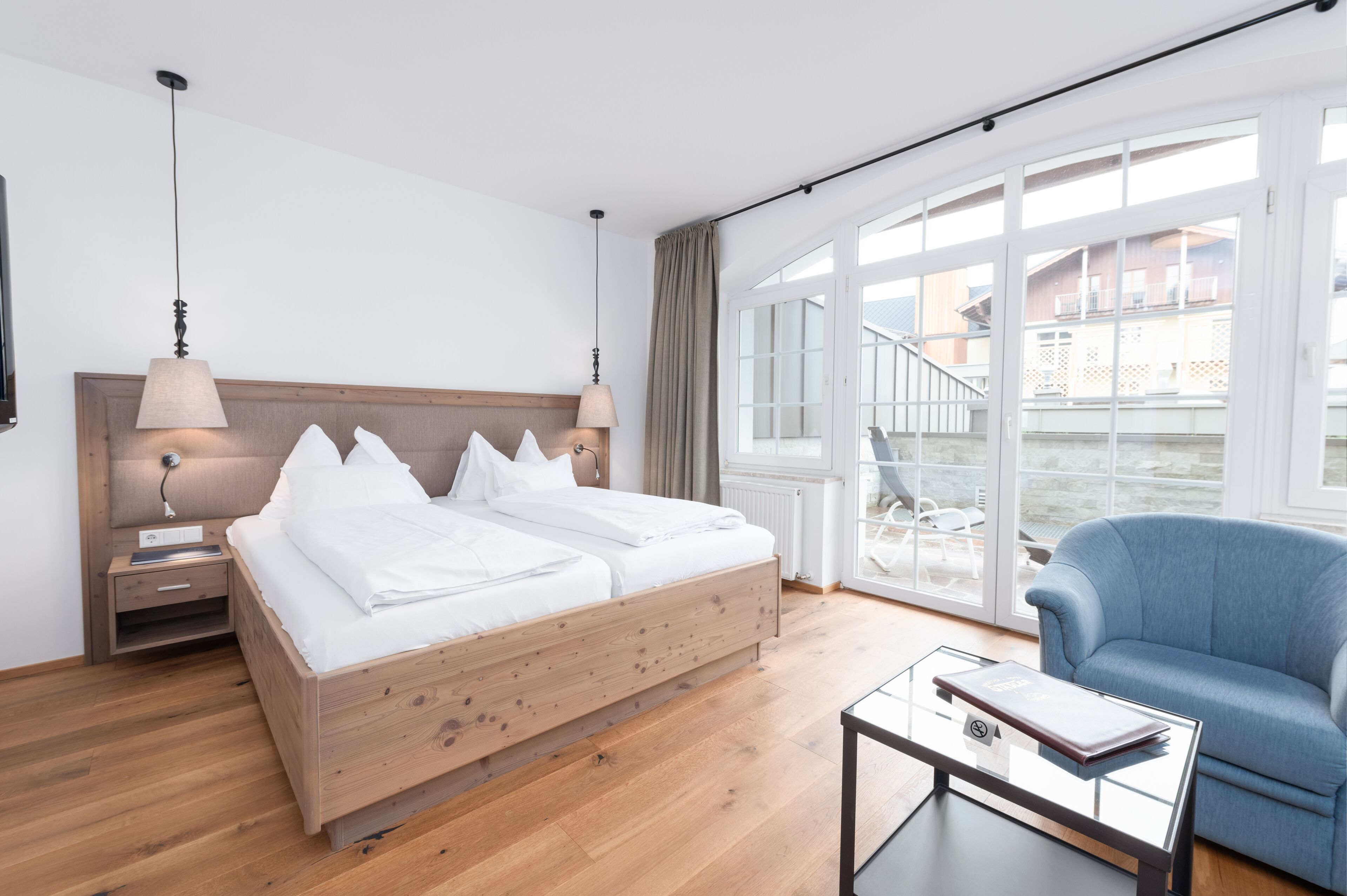 Ein modernes Hotelzimmer mit einem hölzernen Bett, einem Glastisch, einem blauen Sessel und großen Fenstern mit Blick ins Freie.