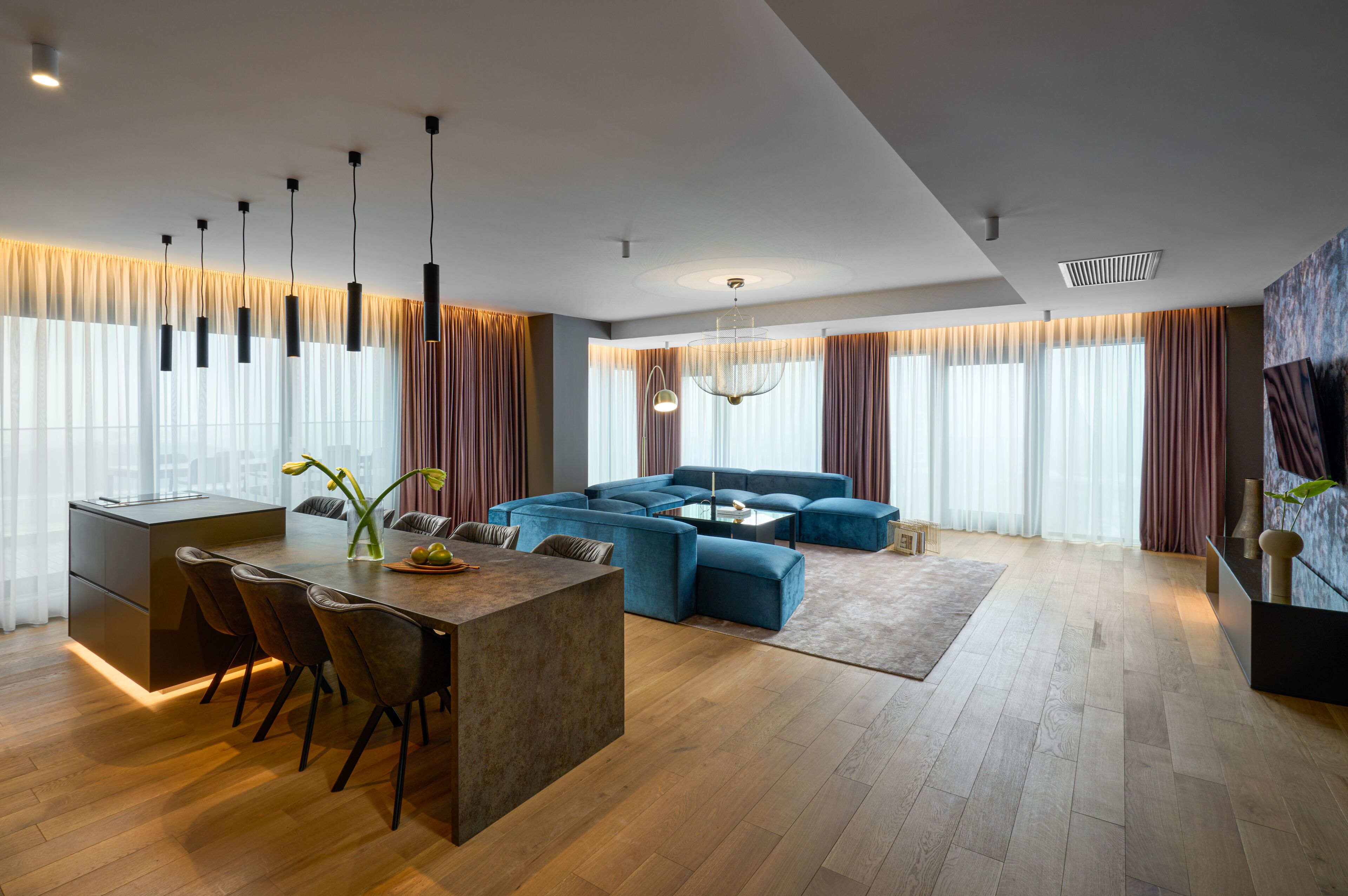 A modern living room with a blue sectional sofa, a dining area with a central island, and a kitchen with a dark countertop.