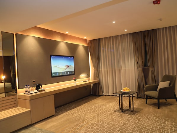 A modern hotel room with a large flat-screen TV mounted on the wall, a wooden desk with a coffee maker, a vase with flowers, a chair, and a patterned carpet.