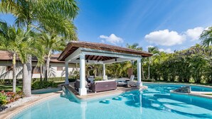 A swimming pool with a covered seating area, palm trees, and a clear blue sky. - Beautiful Villa with Pool Gym and Maid (Punta Cana)