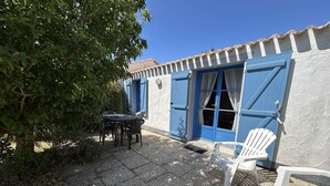 A patio area with a table and chairs, a white plastic chair, and a building with blue shutters and a tiled roof. - Cozy 2-BR House Near Beach with Garden, BBQ, and Terraces - Sleeps 4 (Noirmoutier-en-l'Île)