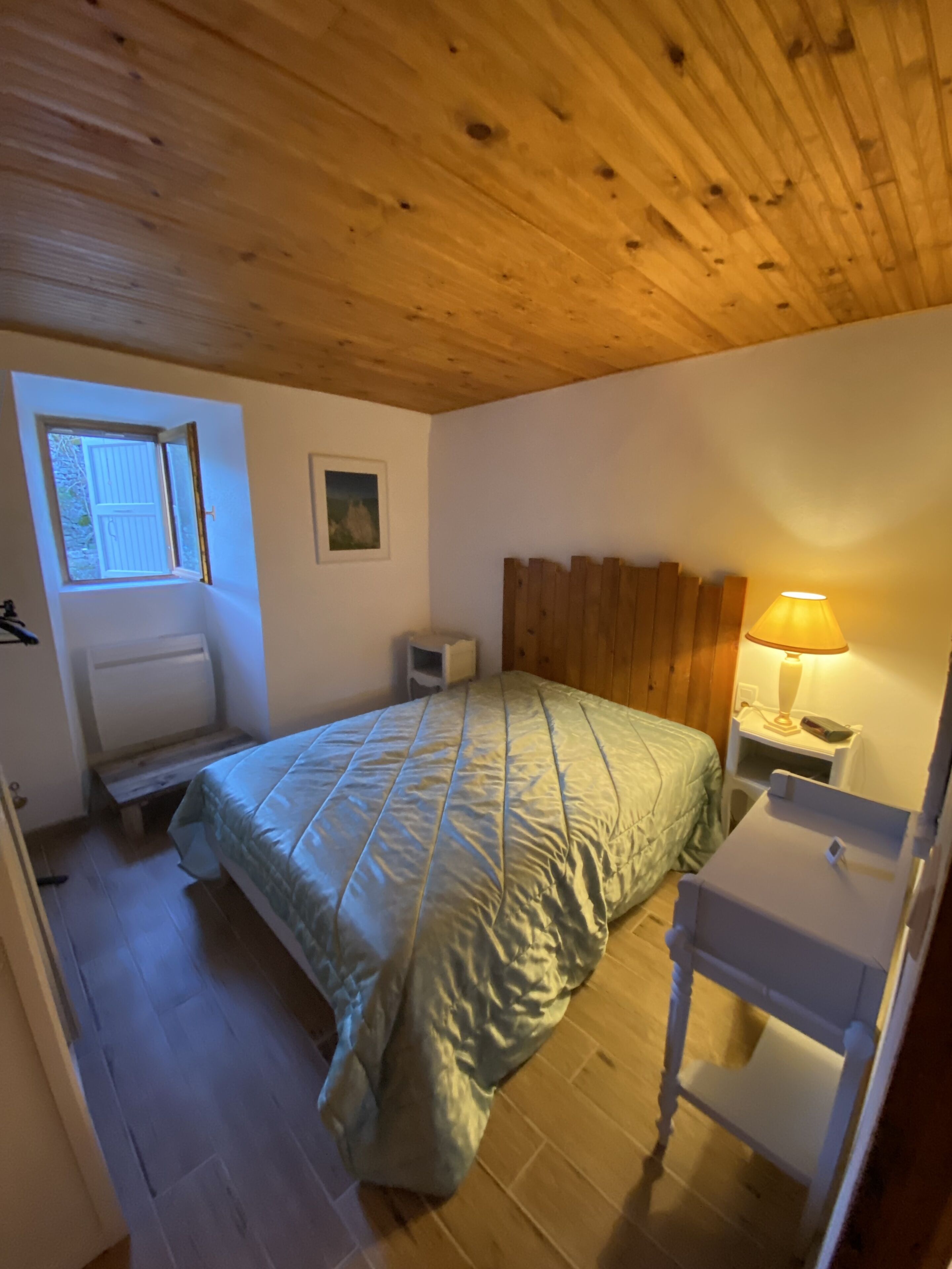 Une chambre à coucher avec un plafond en bois, un lit recouvert d’une couverture verte, une table de chevet avec une lampe et une petite fenêtre.
