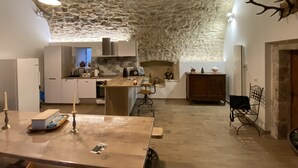 Eine rustikale Küche mit Steinwänden, Holzschränken und einer Steindecke. Ein Holztisch steht dort mit einer Kerze und einem Buch, und eine Bank mit Kleiderhaken.