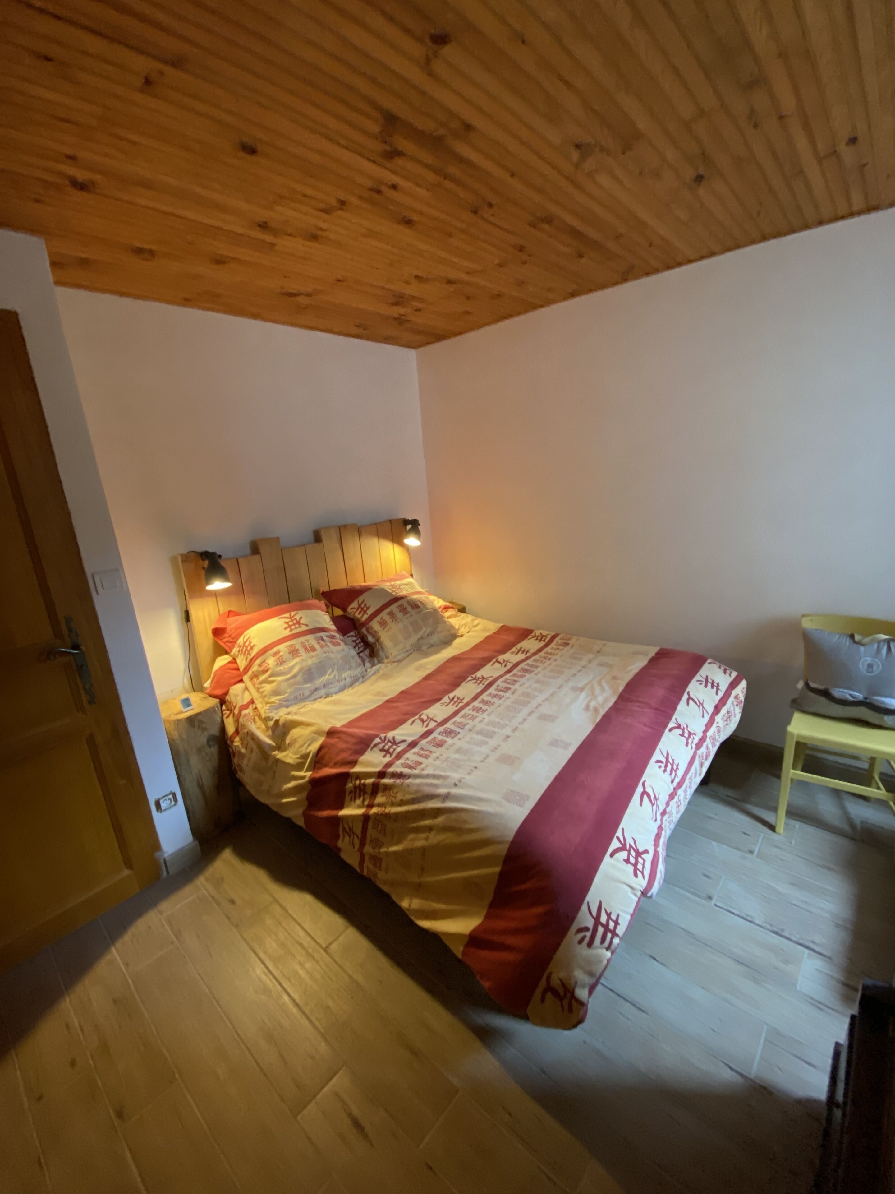 A bedroom with a wooden ceiling, a bed with patterned bedding, two bedside lamps, and a wooden floor.