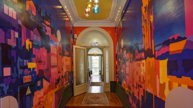 A brightly lit hallway with colorful abstract murals on the walls, a wooden staircase, and a 'Welcome' sign at the end.
