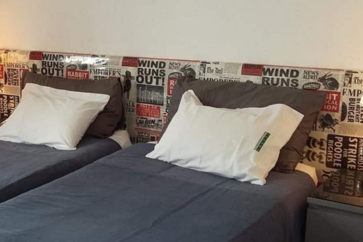 A double bed with two pillows, a red chair, and a wall with newspaper clippings.