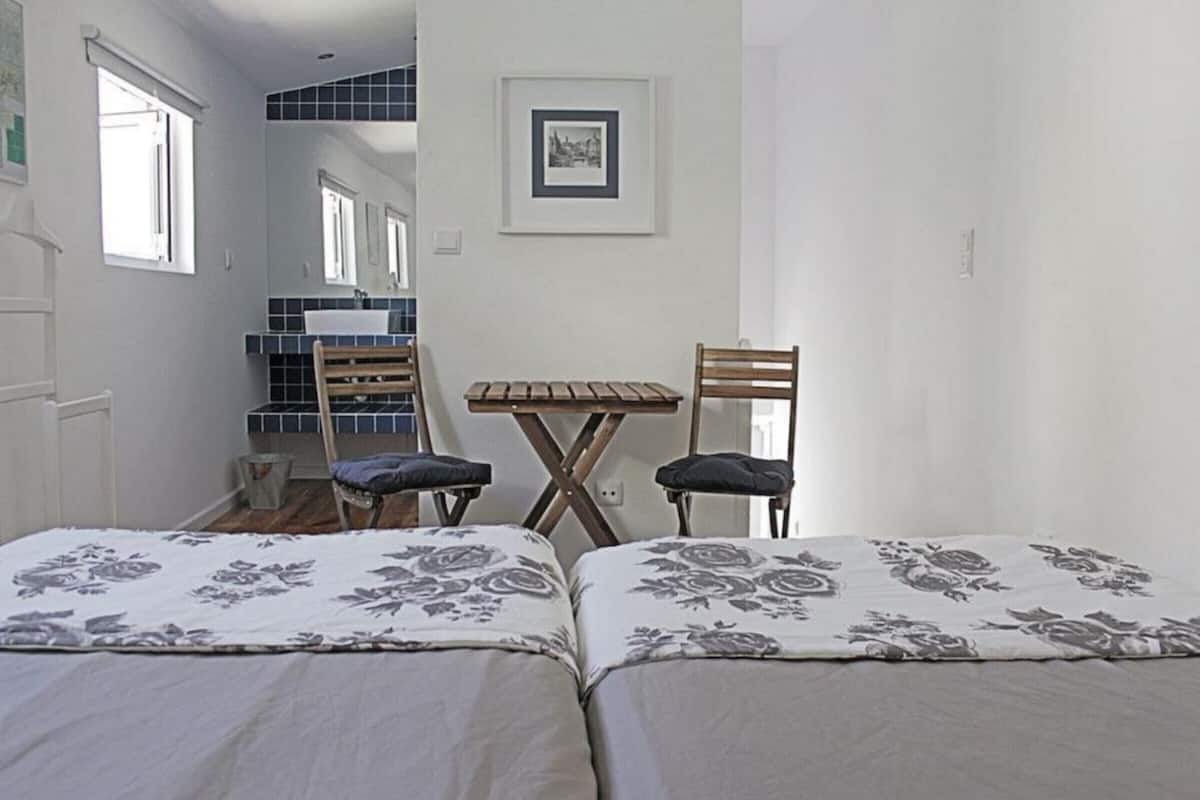 A bedroom with two beds, a small table, two chairs, a window, and a bathroom visible in the background.
