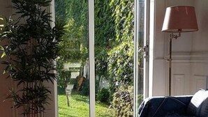 A room with a wooden floor, a black sofa, a potted plant, and a view of a garden through a glass door.