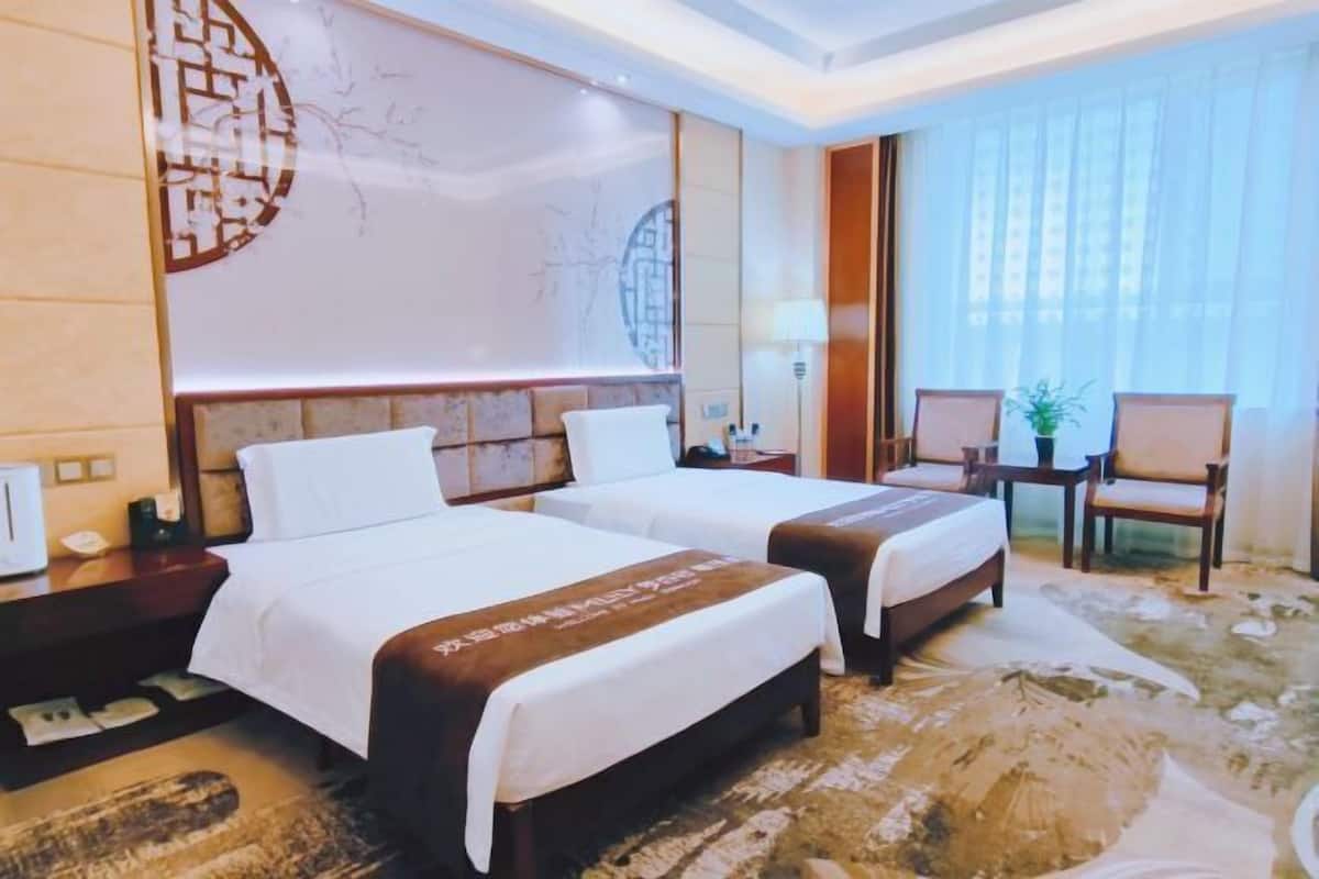 A hotel room with two beds, a nightstand, a lamp, a chair, and a large window with curtains.