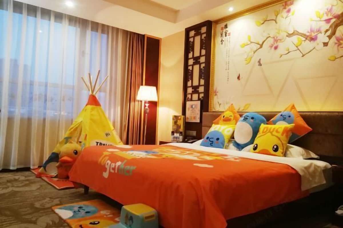 A hotel room with a bed, pillows, a lamp, a window with curtains, and a wall with a floral design.