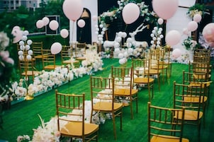 Un espacio con hileras de sillas doradas dispuestas sobre una alfombra verde, rodeado de arreglos florales y globos blancos y rosas.