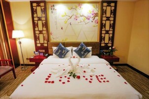 A hotel room with a bed, pillows, a wall with a floral painting, a lamp, and a small table with a vase.