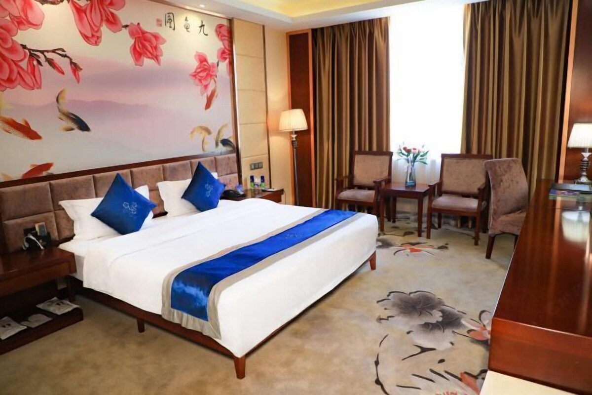 A hotel room with a large bed, a desk, two chairs, a lamp, and a decorative wall with floral patterns.