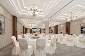 A banquet hall with round tables, white chairs with gold sashes, chandeliers, and framed artwork on the walls.
