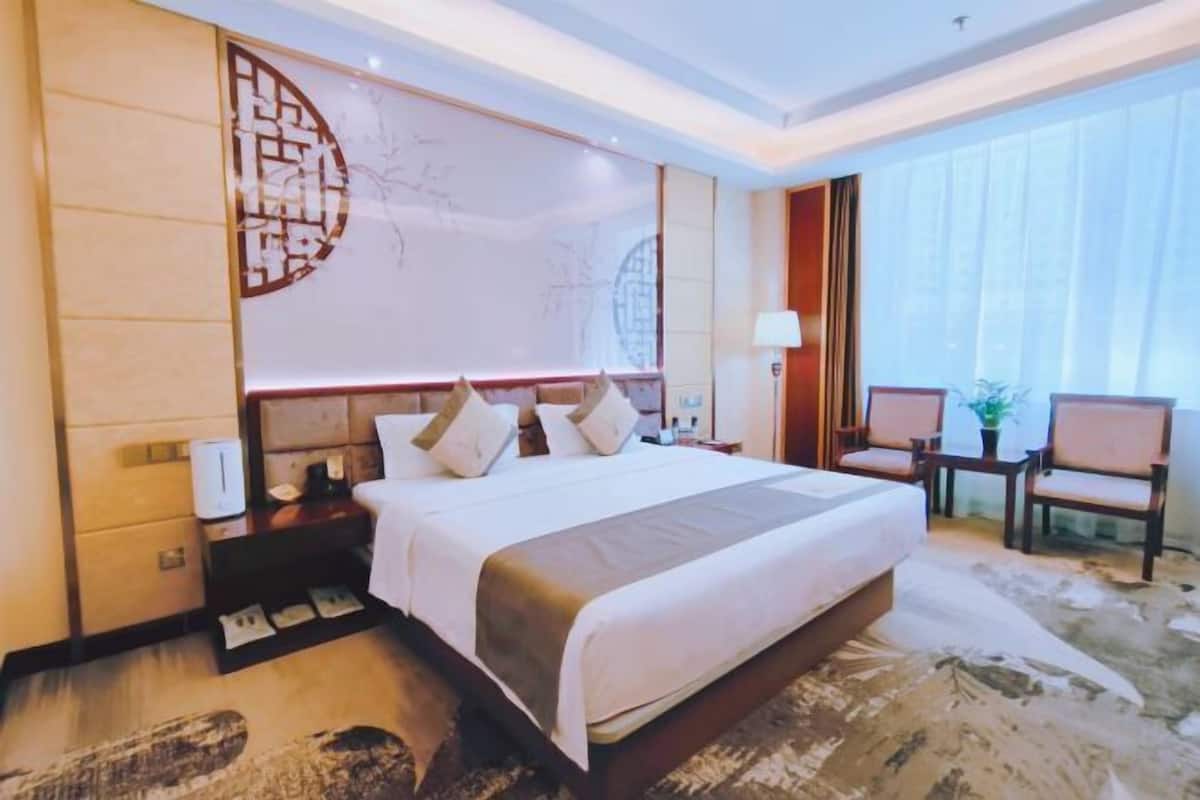 A hotel room with a large bed, bedside tables, a nightstand, a chair, and a large window.