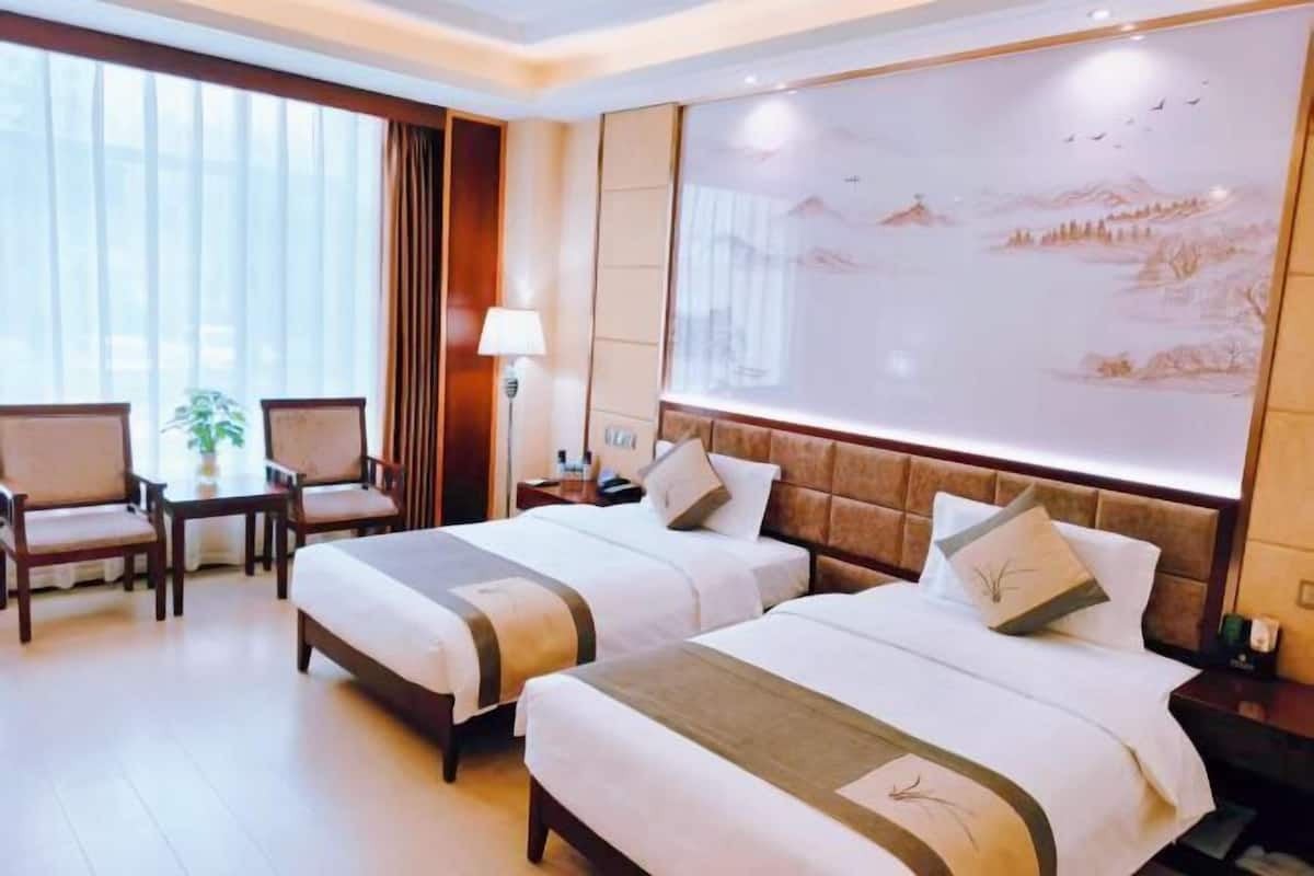 A hotel room with two beds, a desk, chairs, a lamp, and a large wall mural.