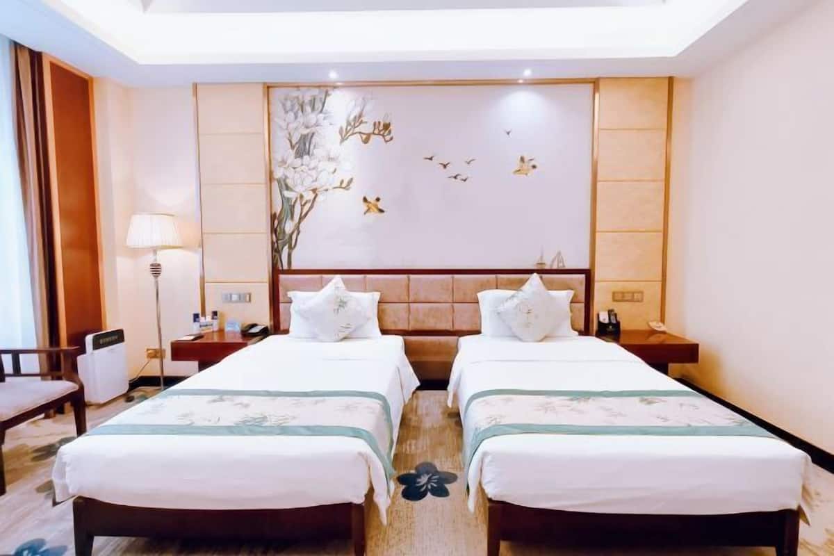 A hotel room with two beds, a nightstand, a lamp, and a wall with a floral painting.