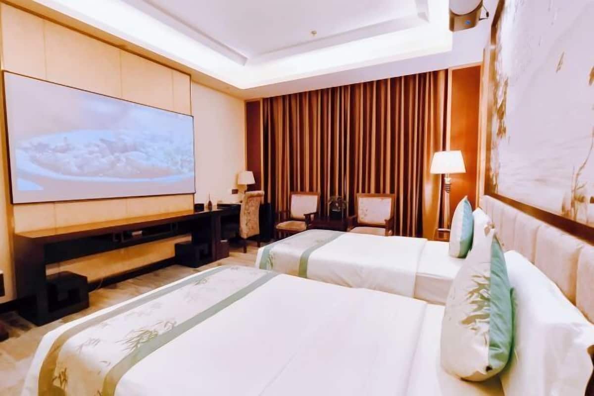 A hotel room with a large flat-screen TV, two beds with white bedding, a wooden panel wall, and a ceiling light fixture.
