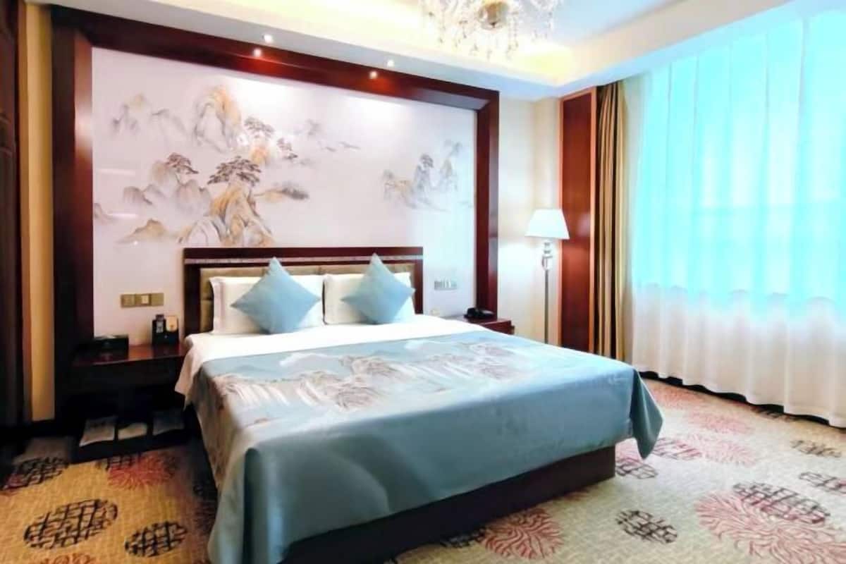 A hotel room with a bed, a nightstand, a wall with artwork, a lamp, and a window with curtains.