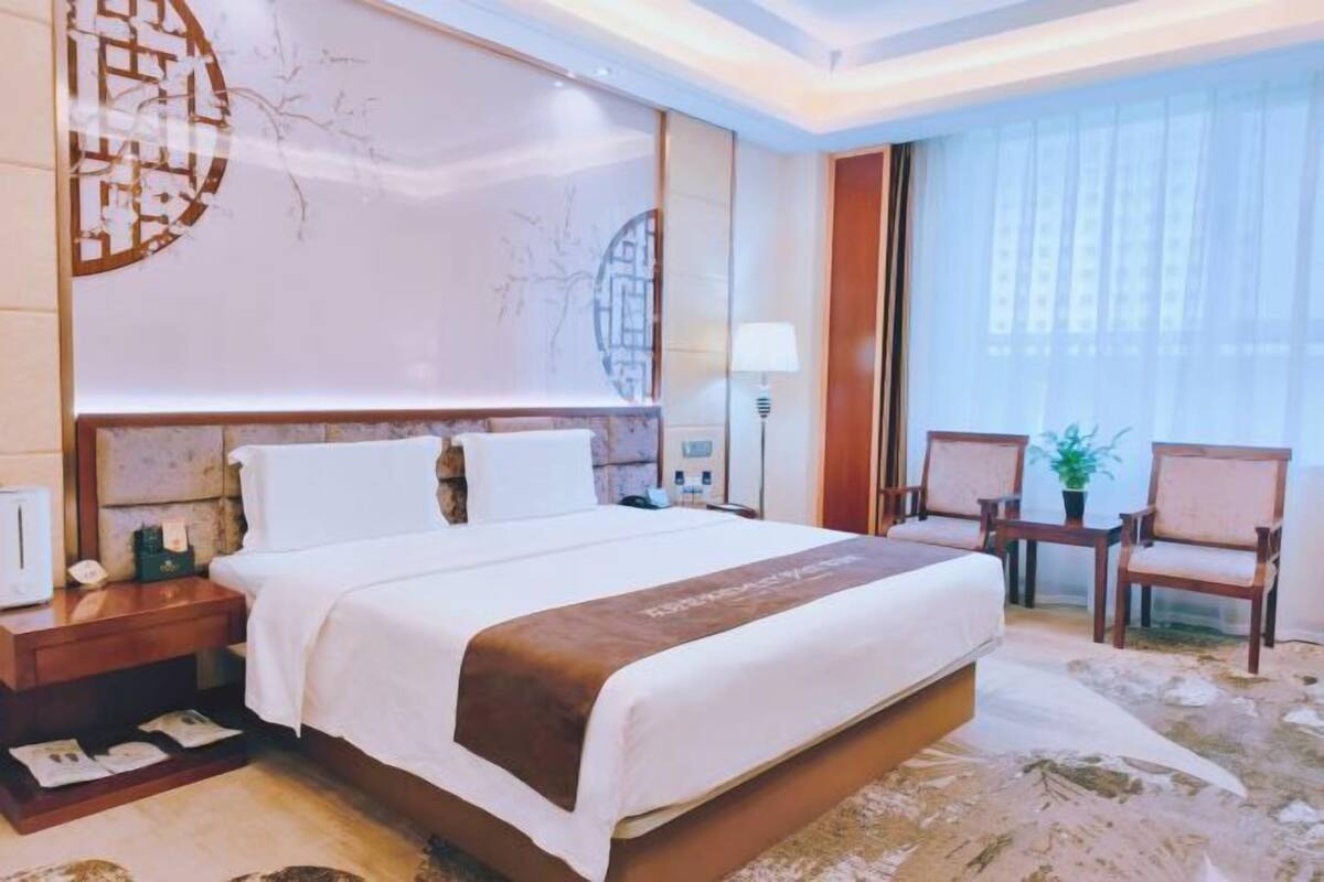 A hotel room with a large bed, bedside tables, a desk, chairs, and a large window with curtains.