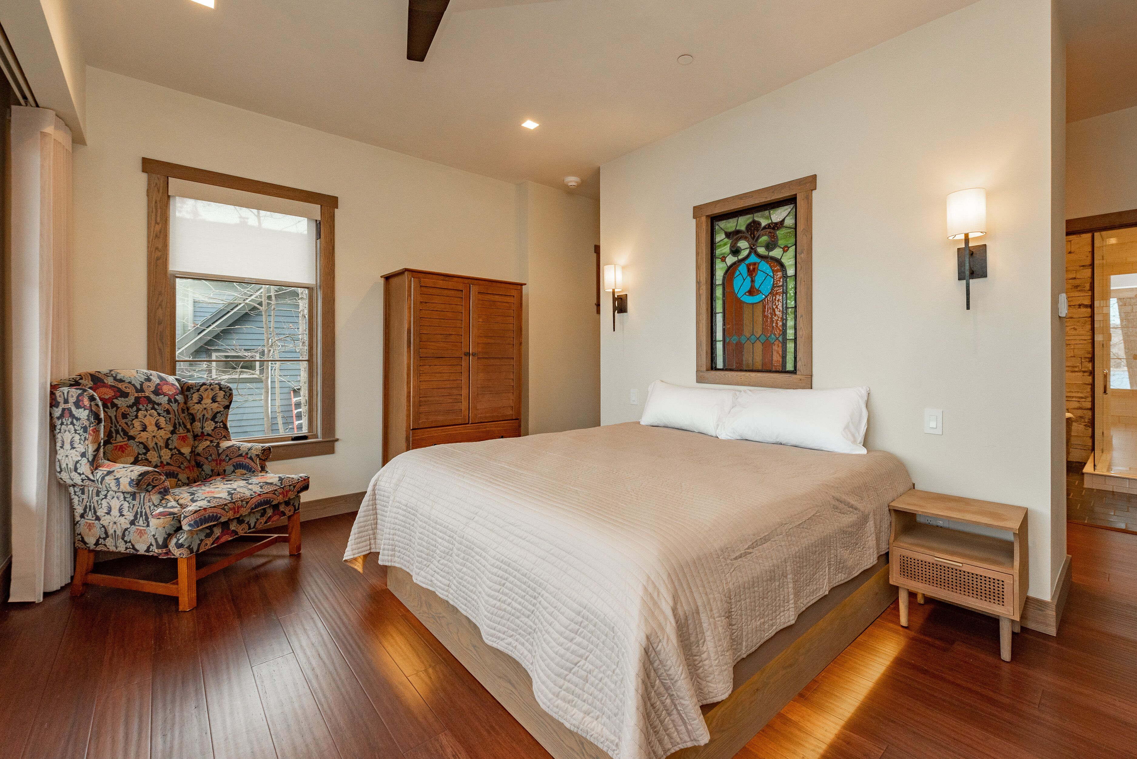 A bedroom with a large bed, a wooden chair, a wooden wardrobe, and a stained glass window.